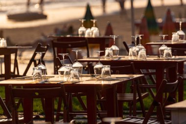 Açık İtalyan kafe veya restoranındaki masalar gün batımında deniz manzaralı kalabalık kumlu plajda akşam yemeği için servis ediliyor.