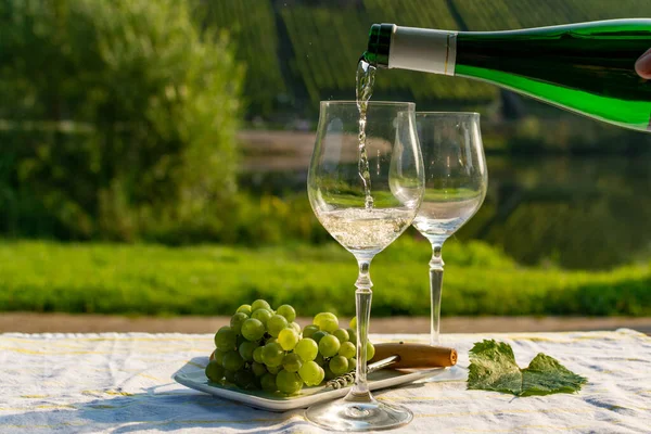 Waiter pouring  German quality white wine riesling, produced in Mosel wine regio from white grapes growing on slopes of hills in Mosel river valley in Germany, bottle and glasses served outside in Mosel valley