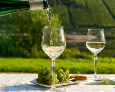 Waiter pouring  German quality white wine riesling, produced in Mosel wine regio from white grapes growing on slopes of hills in Mosel river valley in Germany, bottle and glasses served outside in Mosel valley clipart