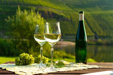 Famous German quality white wine riesling, produced in Mosel wine regio from white grapes growing on slopes of hills in Mosel river valley in Germany, bottle and glasses served outside in Mosel valley clipart