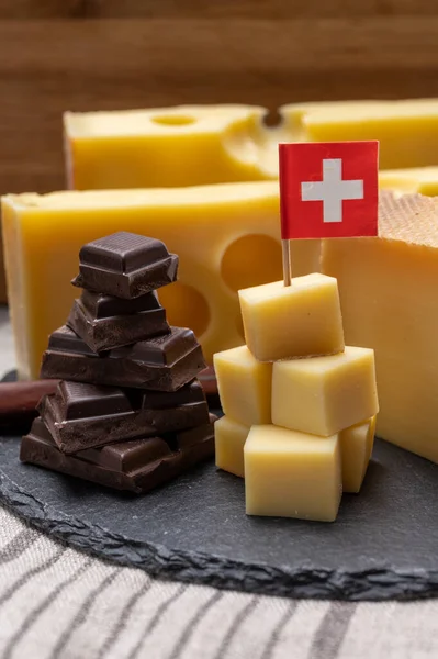 Tasty Swiss food, block of medium-hard yellow cheese emmental or emmentaler with round holes, matured gruyere and high quality milk chocolate close up served in cubes as mountains top with Swiss flag.