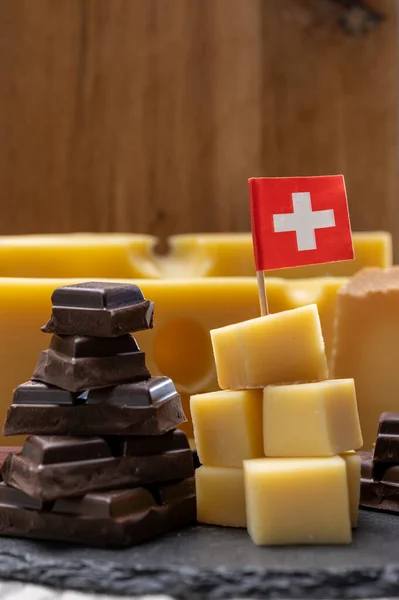 Tasty Swiss food, block of medium-hard yellow cheese emmental or emmentaler with round holes, matured gruyere and high quality milk chocolate close up served in cubes as mountains top with Swiss flag.