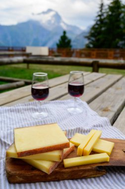 Cheese collection, French comte, beaufort or abondance cow milk cheese and glasses of red wine from Savoie served outdoor with Alps mountains peaks in summer on background clipart