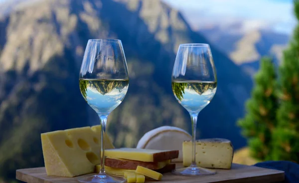 Tasty cheese and wine from Savoy region in France, beaufort, abondance, emmental, tomme and reblochon de savoie cheeses and glass of white wine served outdoor in summer with Alpine mountains peaks on background
