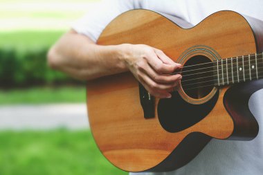 Kahverengi bir klasik akustik gitarın yakın çekimi, erkek bir müzisyenin eli telleri çalıyor. Bulanık doğal bir arkaplan üzerine yakın çekim yapan bir kişinin ellerinde telli müzik aleti.