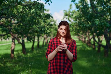 Siyah ve kırmızı tişörtlü genç bir kız kırmızı saçlı yeşil arka planda duruyor ve elinde beyaz bir karahindiba tutuyor, ona bakıp gülümsüyor.
