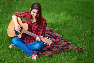Doğada akustik gitar çalan zeki bir kız. Genç bir kadın elinde battaniyeyle yeşil çimlerde oturur ve müzik aleti çalar.