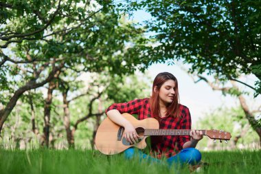 Damalı gömlekli ve kotlu bir kız yeşil çimlerde oturuyor, klasik akustik gitar çalıyor ve kendi elleriyle şarkı söylüyor. Doğada bir kız müzikal bir şeyler çalar.