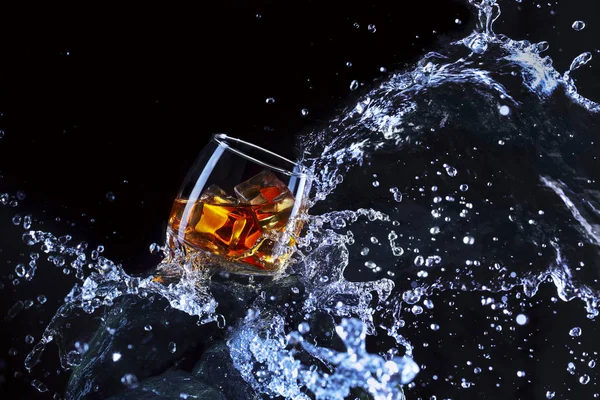 Glass of whiskey with ice .Water splash the glass.Creative photo glass of whiskey on stone with fog and black background.Copy space.Advertising shot — Stock Photo, Image