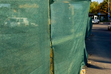 İşçiler, öğleden sonra güneşli bir caddede yeşil ağ çitlerinin arkasında bir inşaat projesini tamamlıyorlar..