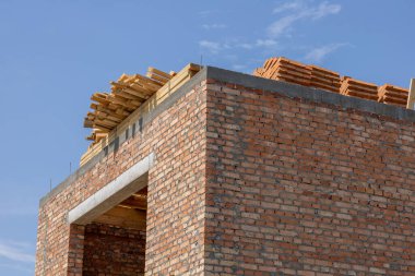 Tamamlanmamış kırmızı tuğla duvarlar ve ahşap çatı çerçevelerini gösteren inşaat projesi. Açık mavi gökyüzünün altında yığılmış yapı malzemeleri.