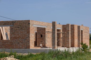 Tamamlanmamış yeni ev inşaatı, kırmızı tuğlalı duvarlar ve parlak mavi gökyüzünün altındaki inşaat sahasının beton temeli..