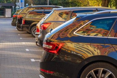 Güneşli bir şehir otoparkında arka arkaya park edilmiş modern arabalar. Koyu renk araç camları ve parlak araba vücutları parlak bina yansımaları.