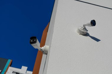 Açık mavi bir gökyüzünün altındaki dokulu bina duvarına monte edilmiş iki beyaz güvenlik kamerası. Kentsel güvenlik için modern izleme teknolojisi.