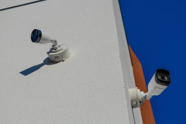 Beyaz güvenlik kamerası ve dış aydınlatma armatürü desenli bir bina duvarına monte edilmiş. Açık mavi gökyüzü, parlak güneş, uzun gölgeler..