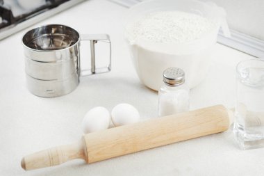 Un, yumurta çırpıcı, merdane, tuz, bir bardak su ve yumurta kabuğu beyazı masa. Mutfak masası un ile ev yapımı yemek yapmak için gerekli malzemelerdir. Taze ekmek ve kek pişirmek için gerekli malzemeler. 