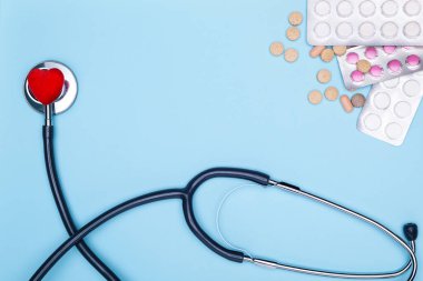 Mavi arka planda bir statoskop, kırmızı bir eyer ve haplar yatıyor. Kardiyovasküler hastalıkların tedavisi. Mavi arka planda boyalı kalp, bir sürü uyuşturucu ve steteskop..