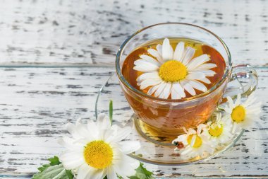 Bir fincan çay ve çay tabağı, ahşap, beyaz bir masanın üzerinde, açık havada, güneş ışınlarında ve papatya çiçeğinin beyaz çiçeklerinde.