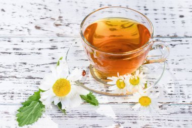 Bir fincan çay ve çay tabağı, ahşap, beyaz bir masanın üzerinde, açık havada, güneş ışınlarında ve papatya çiçeğinin beyaz çiçeklerinde.