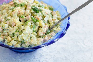 Beyaz bir masa örtüsü üzerinde mavi tabakta zeytinli Rus salatası..