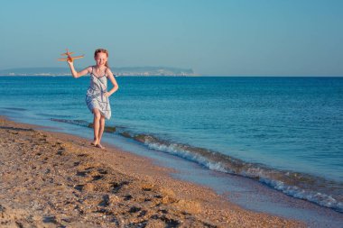 Güzel kızıl saçlı kız, oyuncak bir uçakla oynuyor, açık mavi bir gökyüzünün arka planında denizin kumlu sahilinde koşuyor ve güneş ışınlarından ve özgürlükten zevk alıyor.