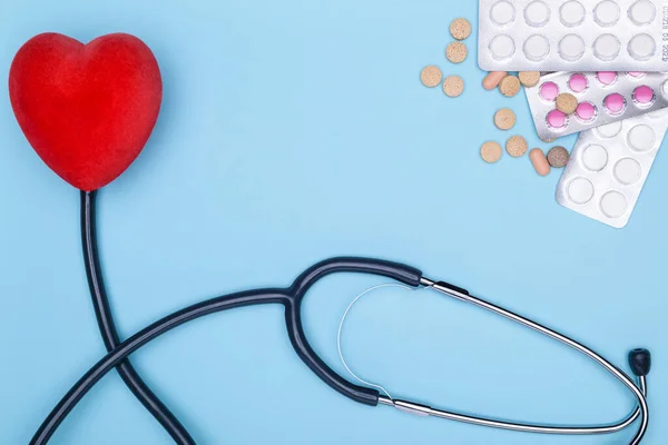Concept of treatment of cardiovascular diseases. Closeup of painted heart, lot of drugs and stethoscope on blue background.Blue background lies a statoscope, a red saddle and pills.