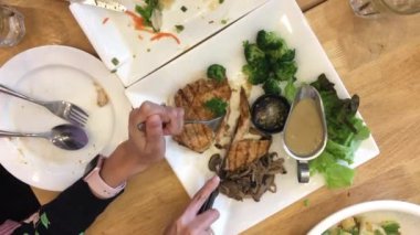 Kadın elinde bıçak ve çatalla ızgara bifteği yemek setiyle birlikte beyaz tabakta keser ve diğeri de restorandaki ahşap masada bitirir. Üst Manzara. video görüntüleri.