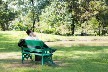 Yeşil Ahşap bankta oturan ve bir beyaz kağıt bardak su, kahve veya çay için elinde tutan genç iş adamı almak uzakta ve içme, yeşil orman park açık havada çalıştıktan sonra rahatlatıcı.