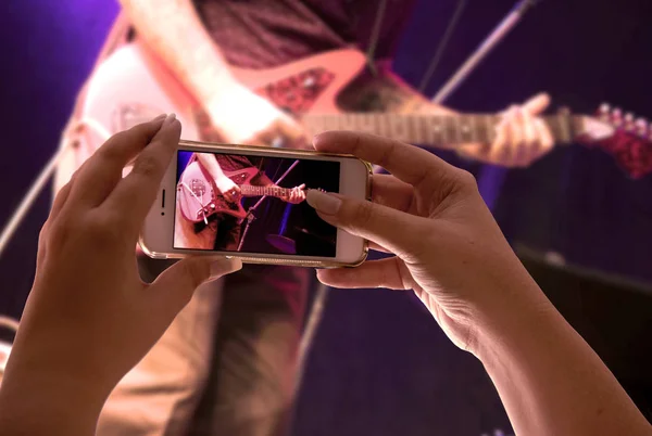 woman\'s hand holding a smartphone in audience recording a musical presentation with electric guitar close up on screen
