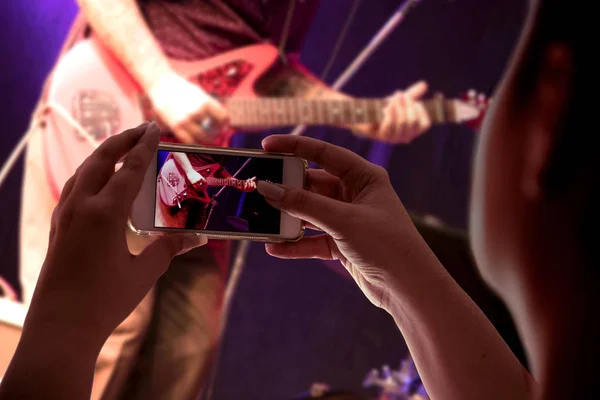 woman\'s hand holding a smartphone in audience recording a musical presentation with electric guitar close up on screen