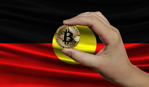 Hand of a man with a gold bitcone Cryptocurrency Digital bit of coins in a hand on a background of the flag of Australian Aboriginal. The concept of virtual money.