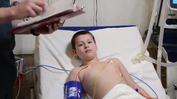 Little boy lying on hospital bed and smiling while listening to male doctor writing on clipboard — Stock Video