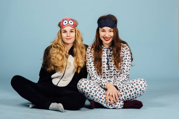 Two women in pajamas sit on floor on a blue background. Studio shoot