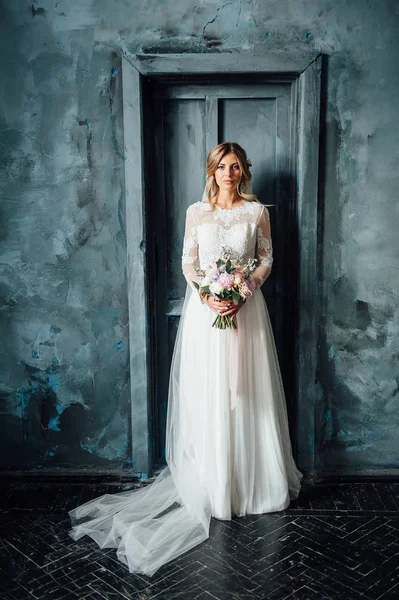 Hermosa novia en vestido de novia blanco con un ramo de boda de pie cerca de la puerta de madera —  Fotos de Stock