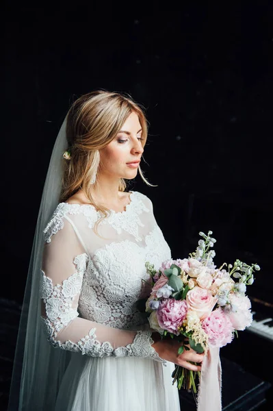 Hermosa novia en vestido de novia blanco con un ramo de boda de pie cerca de la puerta de madera — Foto de Stock