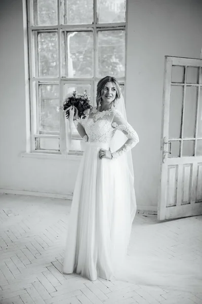 Hermosa novia en vestido de novia blanco con un ramo de boda de pie cerca de la puerta de madera — Foto de Stock