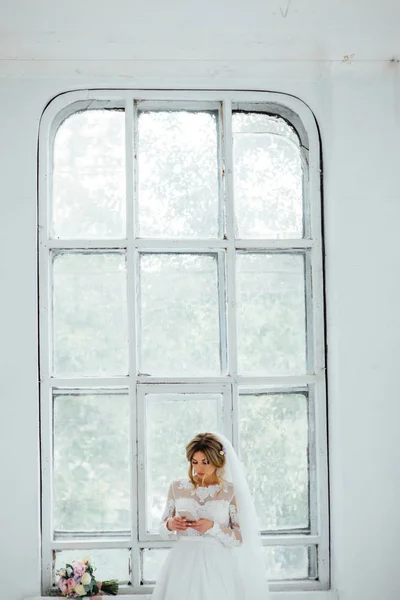 Hermosa novia en vestido de novia blanco con un ramo de boda de pie cerca de la puerta de madera — Foto de Stock