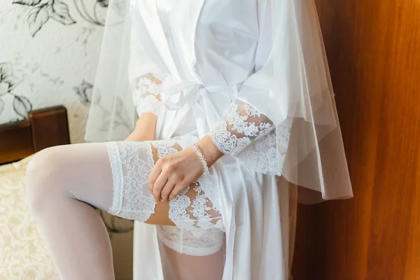 Beautiful bride in white lingerie sitting on bed in her bedroom and putting on stockings — Stock Photo, Image