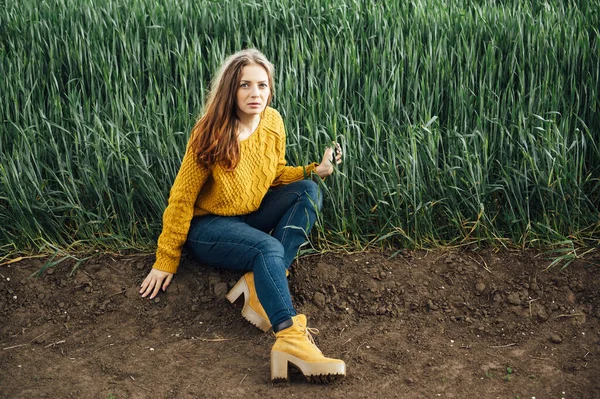 Красивая девушка в джинсах, желтый свитер и сапоги сидит рядом с ранним пшеничным полем — стоковое фото