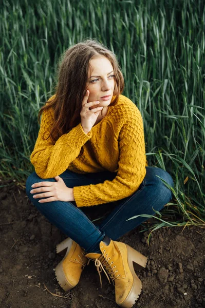 Menina bonita com jeans, uma camisola amarela e botas fica perto de um campo de trigo cedo — Fotografia de Stock