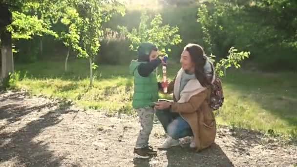 Sorrindo jovem mãe e pequeno filho soprando bolhas sabão juntos em um parque de verão — Vídeo de Stock