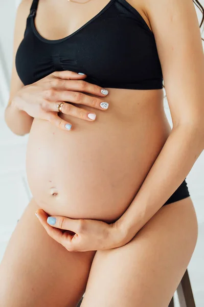 Eine Schwangere mit perfekter Figur sitzt auf einem Stuhl und hält ihren Bauch mit den Händen — Stockfoto