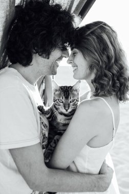 Kumsalda kediye sarılan romantik bir çift. Arka planda gri bir kumaş var.