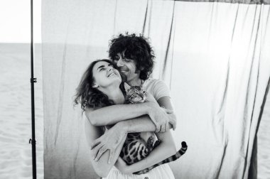 Kumsalda kediye sarılan romantik bir çift. Arka planda gri bir kumaş var.