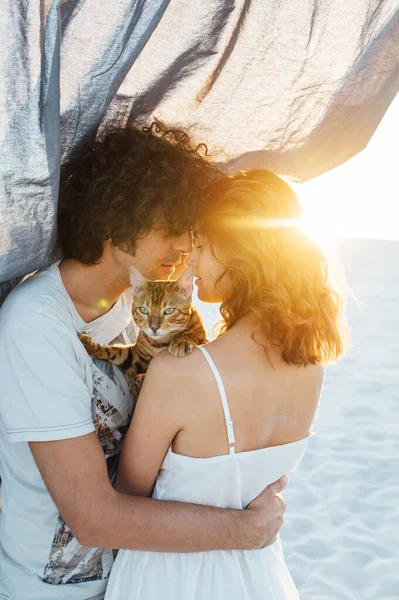 Casal romântico abraçando com um gato na praia. No fundo é um pano cinza — Fotografia de Stock