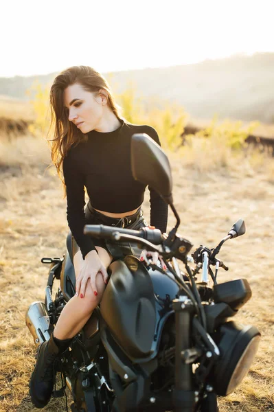 Menina bonita em roupas pretas senta-se na motocicleta e posa para câmera. Pôr-do-sol — Fotografia de Stock