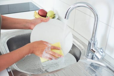 Çekici bir genç kadının kırpılmış görüntüsü evde temizlik yaparken bulaşıkları yıkıyor.