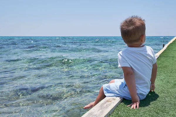 Μικρά παιδιά, κάθεται στην ξύλινη προβλήτα στο νερό και απολαμβάνοντας την καλοκαιρινή μέρα. Γυμνά πόδια του αγοριού. Διακοπές στη θάλασσα. Σε εξωτερικούς χώρους. Τα αδέλφια. Αδελφή και ο αδελφός από τον ωκεανό. — Φωτογραφία Αρχείου