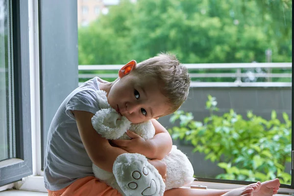Хлопчик два роки сидить біля вікна і обіймає іграшку Кролика. дощова погода, чекає, поки тато повернеться додому з роботи — стокове фото
