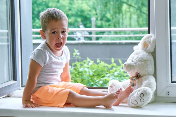 Pojke på två år sitter vid fönstret och kramar en leksak Bunny. regnigt väder, väntar på pappa att komma hem från jobbet — Stockfoto